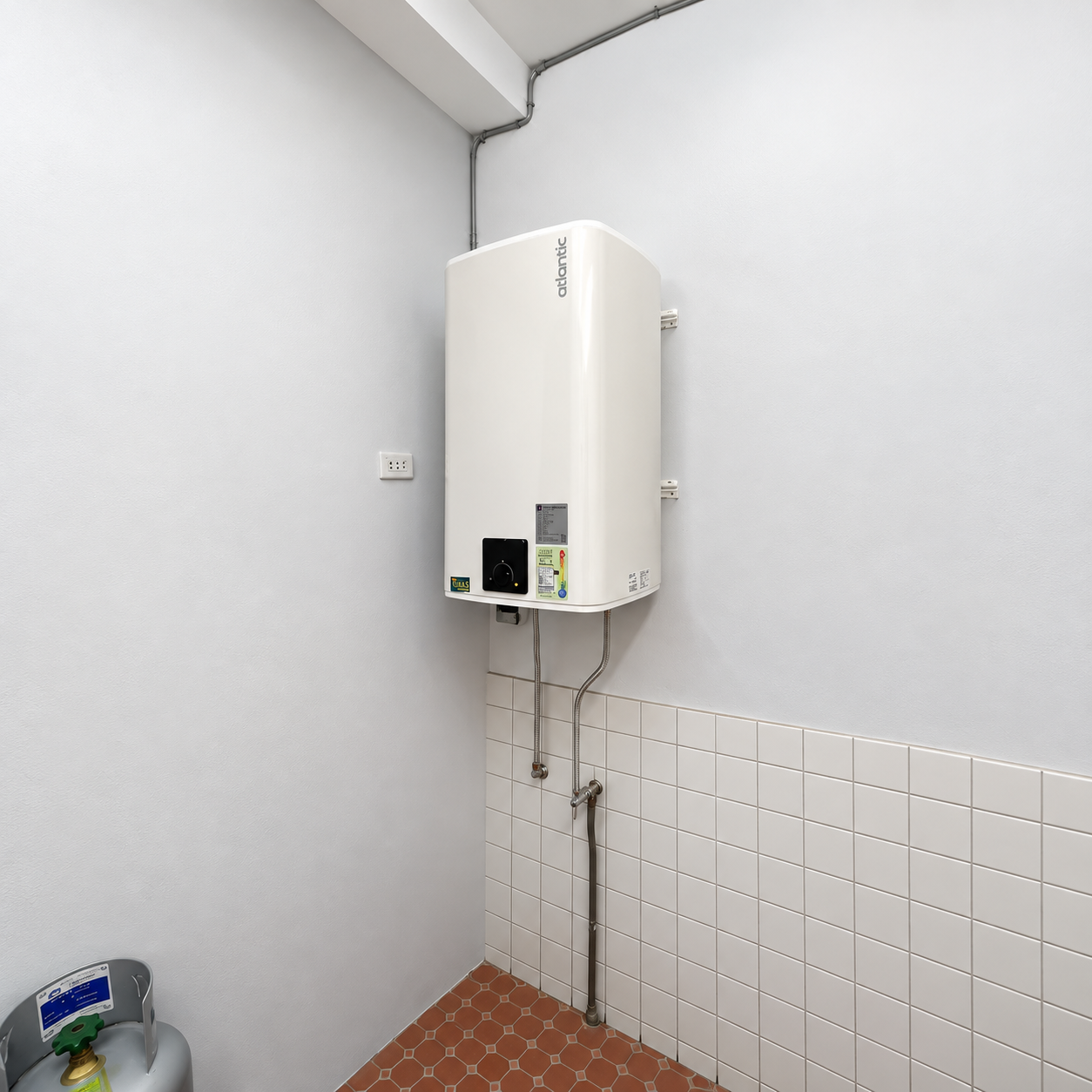 白色方形電熱水器直立壁掛於狹長的磁磚牆邊空間,機身下方可見連接管線與橘色六角地磚。